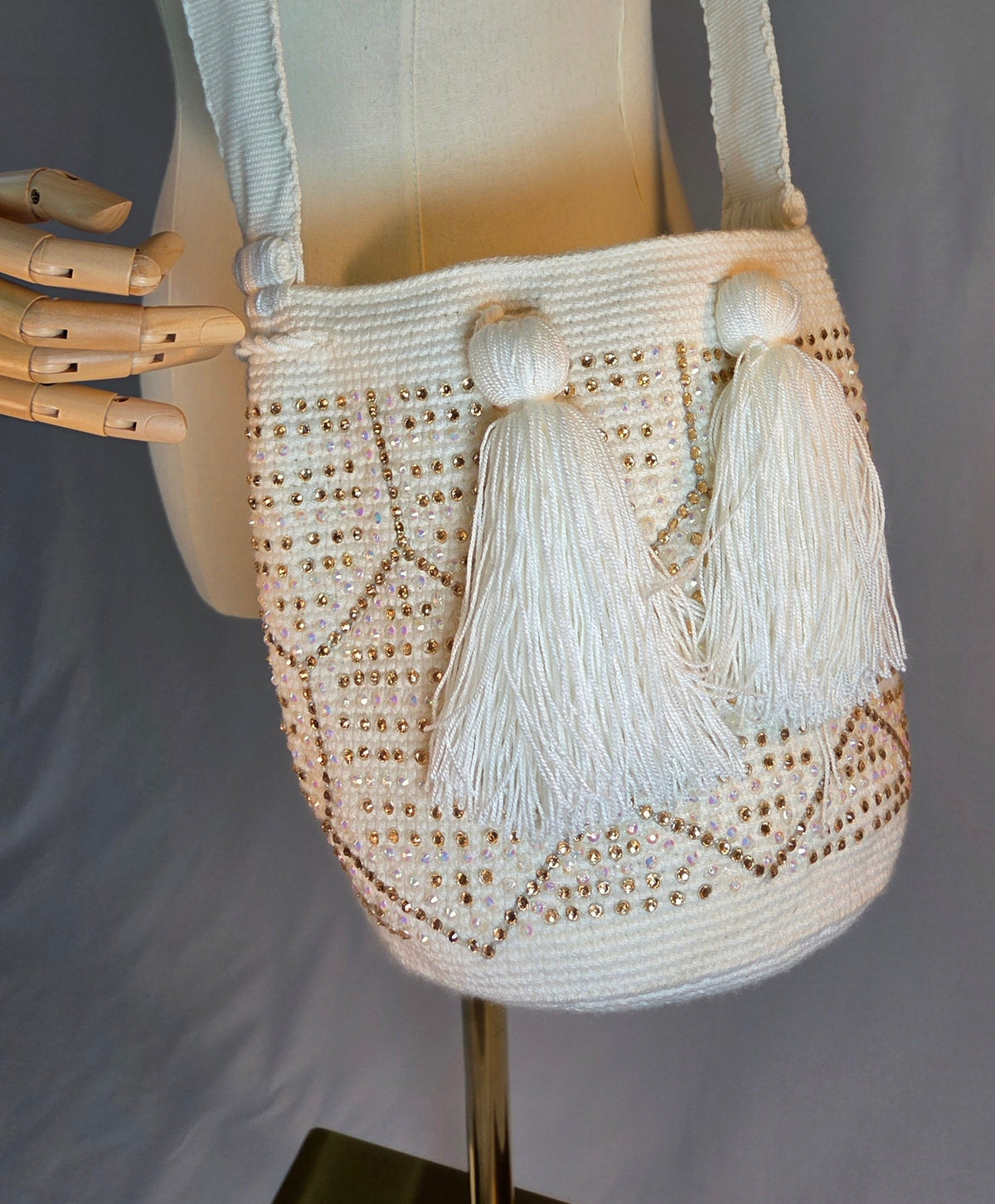A cream-colored crocheted shoulder bag with two large tassel details, displayed on a cream fabric mannequin torso.