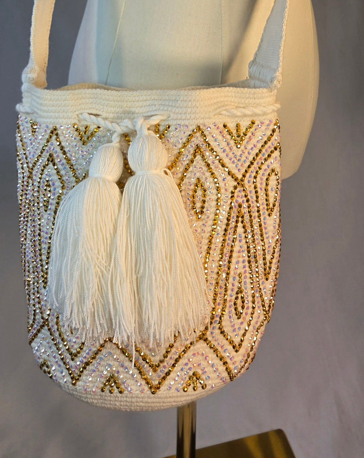 A white woven shoulder bag with gold geometric embroidery and two large white tassels, displayed on a cream-colored mannequin torso with wooden arms against a gray studio background.
