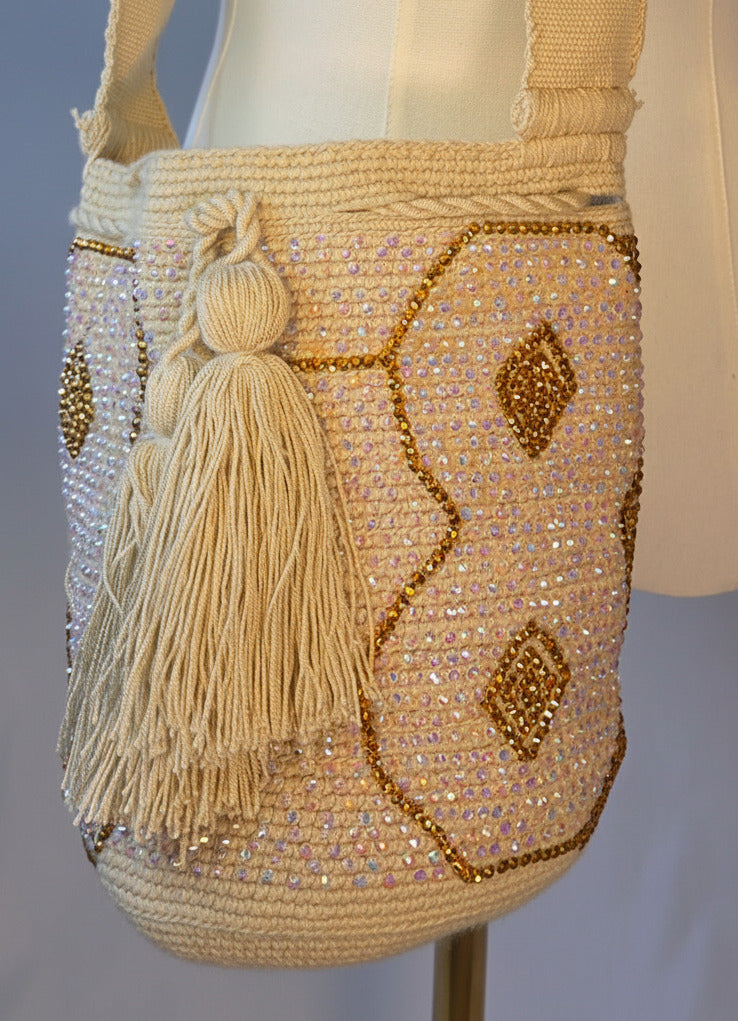 A cream-colored beaded shoulder bag with gold-toned geometric patterns and a tassel, displayed on a cream mannequin torso with wooden arms.